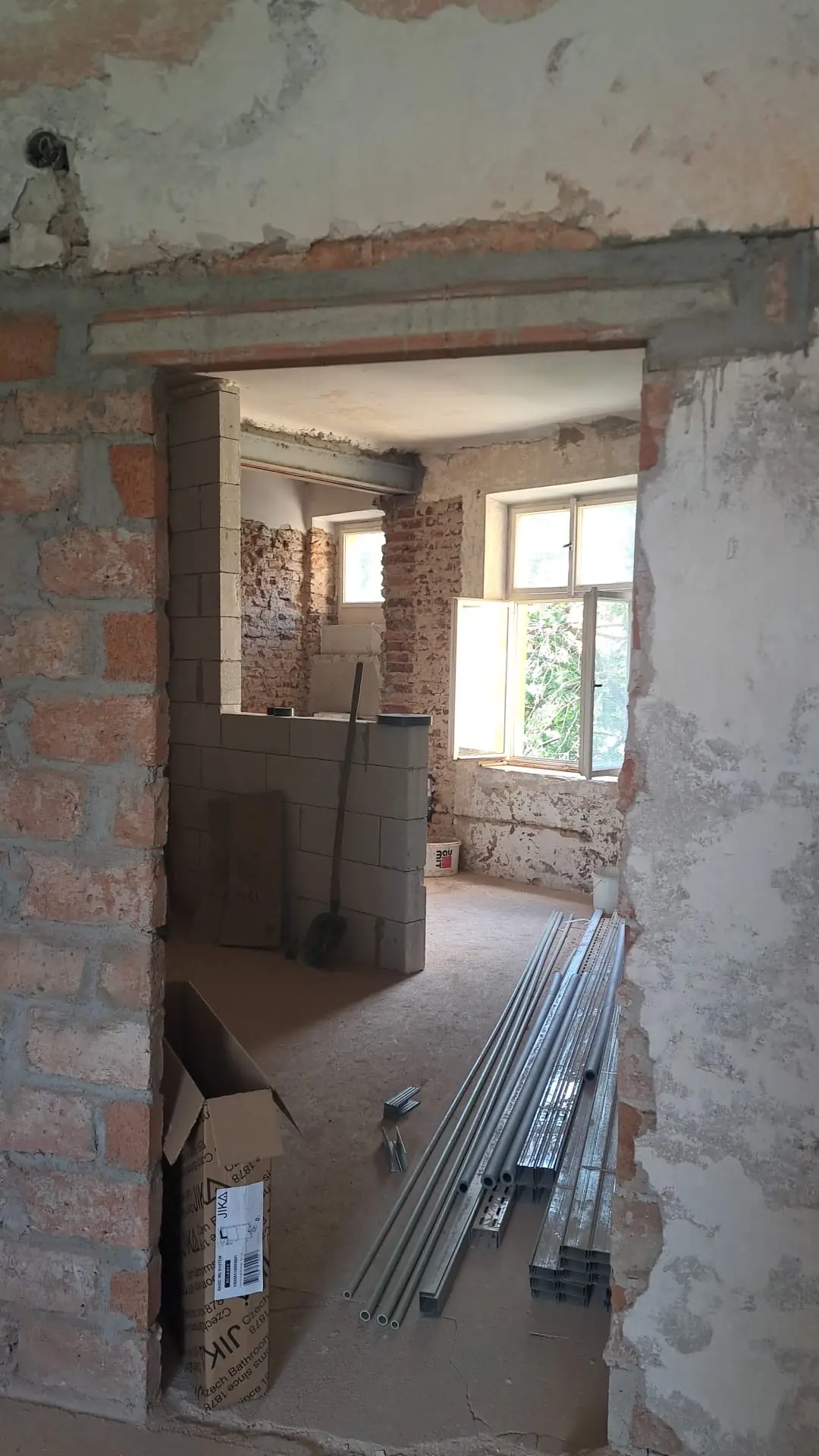 Interiér rozestavěného bytu projektu Bytový dům Otakarova s odhaleným cihlovým a tvárnicovým zdivem, oknem a stavebním materiálem na podlaze.