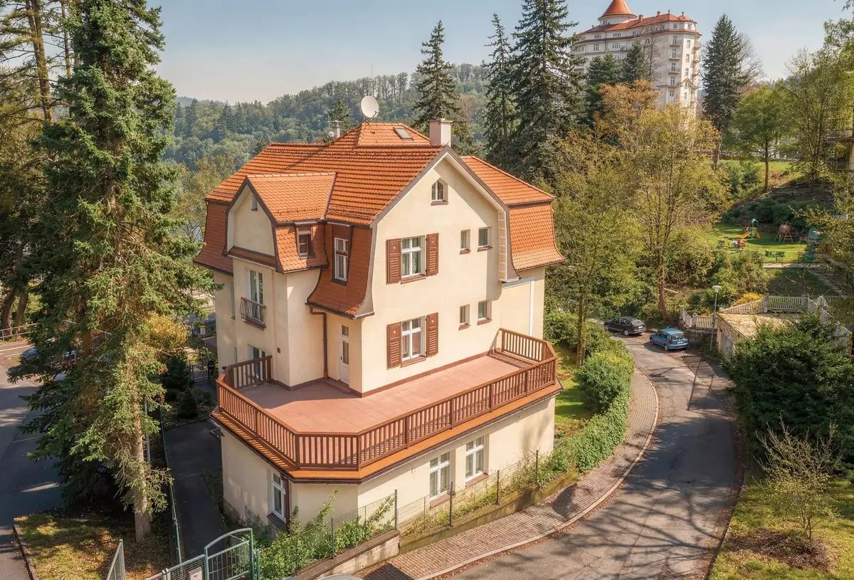 Pětipodlažní historická vila se světlou fasádou a oranžovou střechou v zeleni Karlových Varů, v pozadí hotel Imperial.